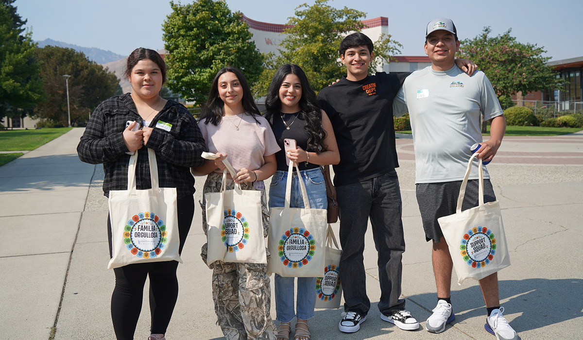 A group of students stand together in front of Van Tassell Center and hold their orientation bags.