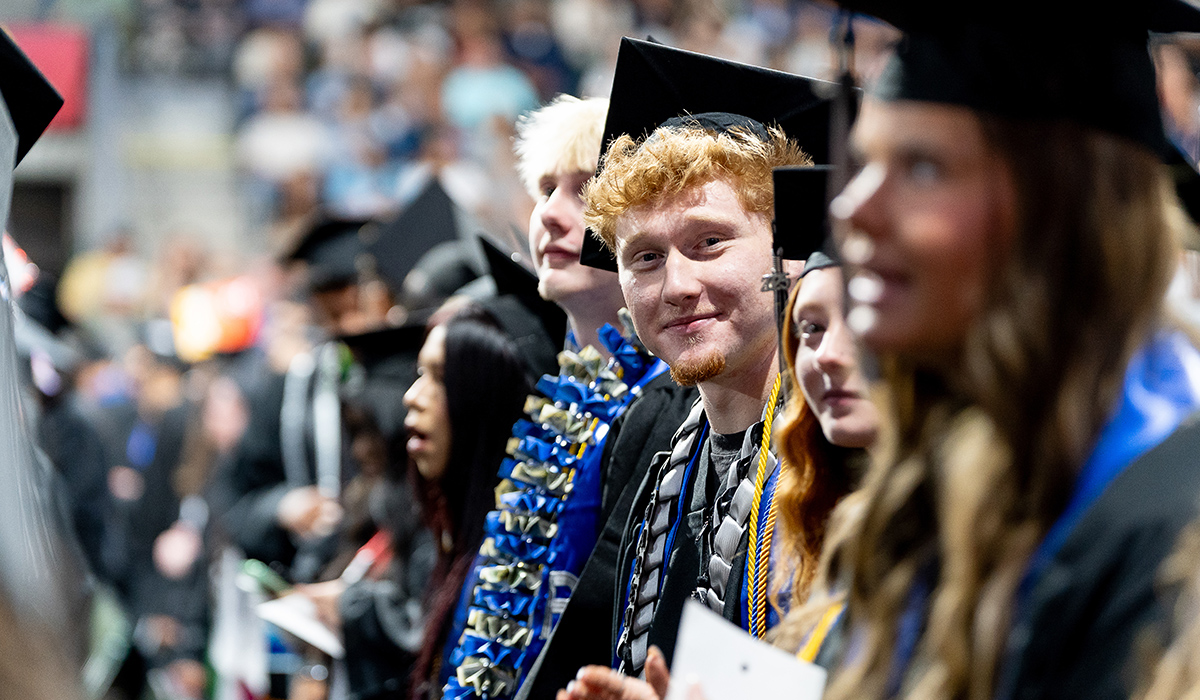 A WVC graduate wearing a black cap and gown sits among other graduates and smiles at the camera.