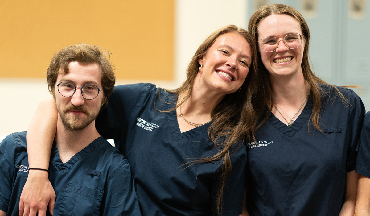 Three nursing students in dark blue scrubs stand together and smile at the camera.