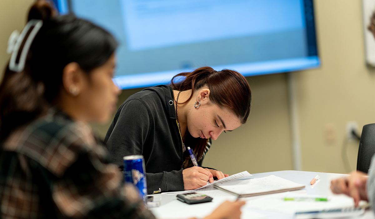 A student writes on a sheet of paper while another student talks with another person.