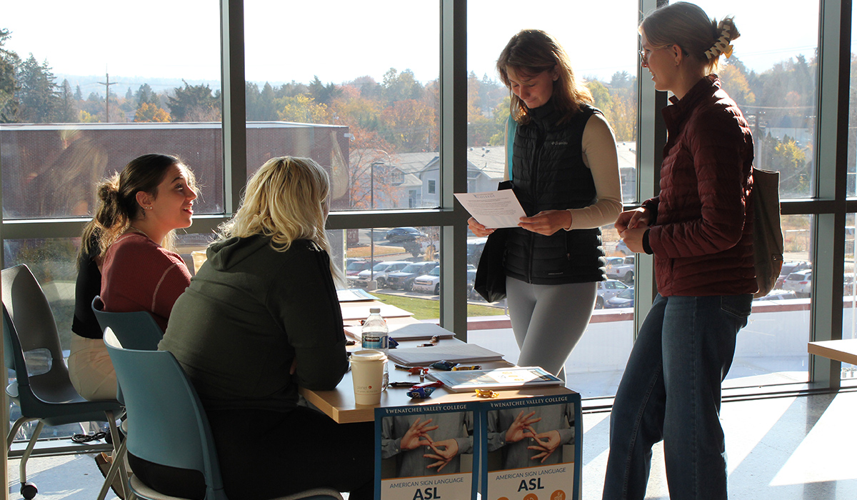 Two students in the ASL program sit at a table at an event and talk with two other students.