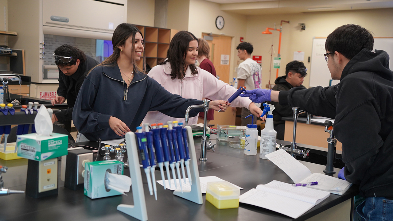 One student passes another student a science tool in a science lab.