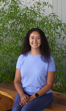 Portrait of Aracely Mendoza sitting on a wooden bench next to a large plant.