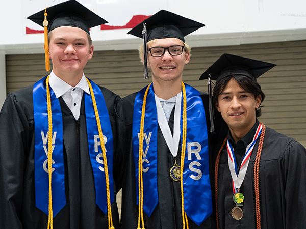Three WVC Omak graduates wearing black robes, blue stoles, and honor cords smile at the camera.